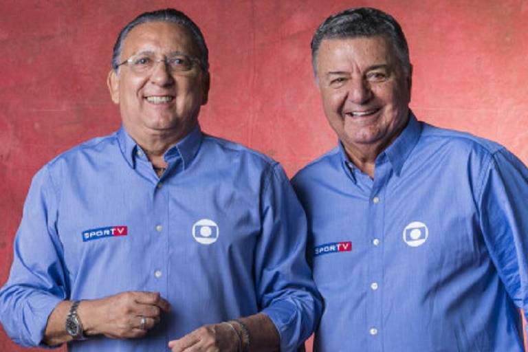 Dois homens sorrindo, vestindo camisas azuis com o logotipo da SporTV. O fundo é de uma cor vermelha suave.