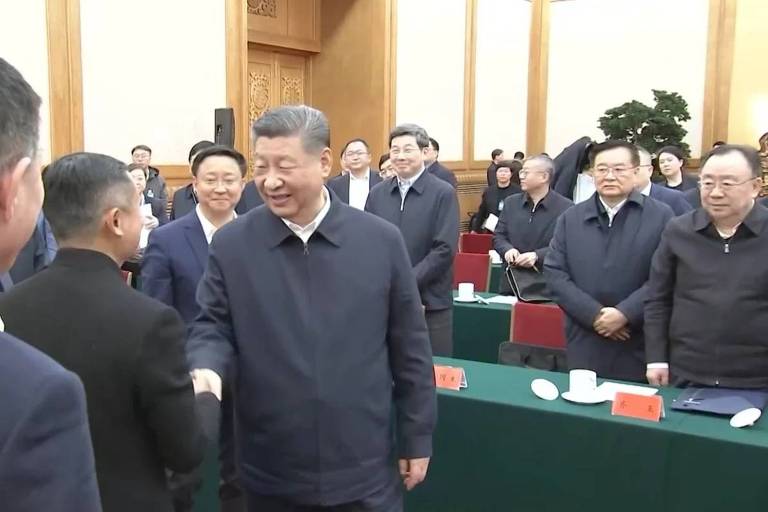 A imagem mostra uma reunião com várias pessoas em um ambiente formal. No centro， um homem de terno escuro está apertando a mão de outro homem， que está vestido com um casaco claro. Ao fundo， há várias outras pessoas observando， algumas sorrindo. O ambiente é bem iluminado， com mesas cobertas por toalhas verdes e cadeiras vermelhas.