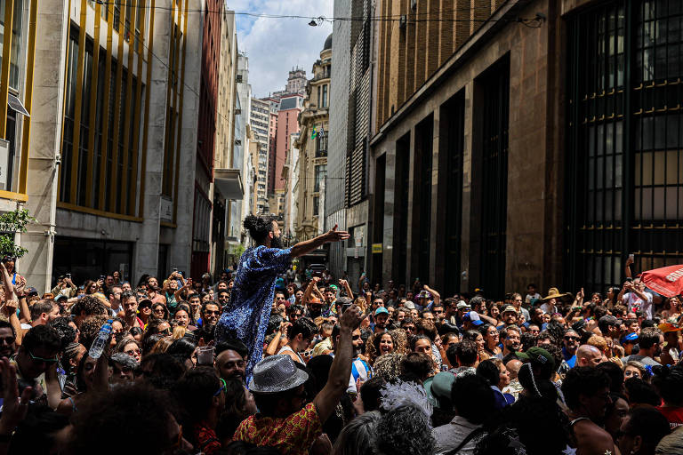 A imagem mostra uma grande multidão reunida em uma rua urbana, cercada por prédios altos. No centro, uma pessoa se destaca, levantando os braços e interagindo com a multidão. A atmosfera parece festiva, com muitas pessoas sorrindo e se divertindo. A iluminação é natural, sugerindo que a cena ocorre durante o dia.