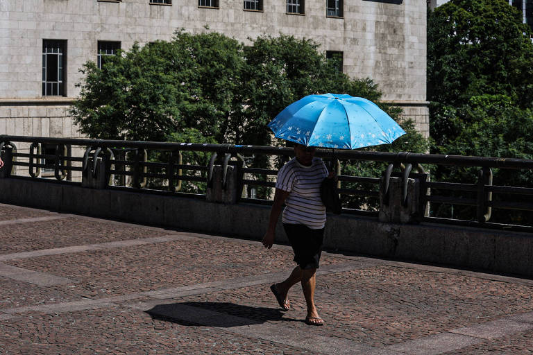 Uma pessoa caminha em uma calçada， segurando um guarda-chuva azul. A pessoa está vestindo uma camiseta listrada e shorts. Ao fundo， há um edifício de pedra e árvores. A luz do sol está forte， criando sombras no chão.