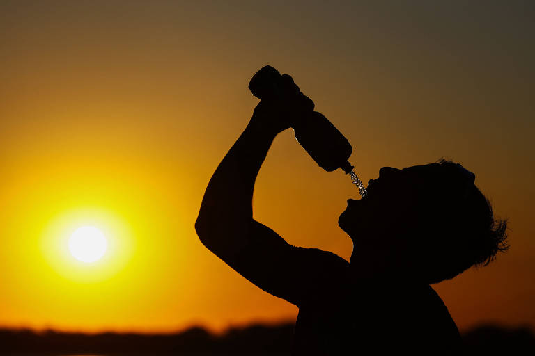 A imagem mostra a silhueta de uma pessoa segurando uma garrafa de água e bebendo， com o sol se pondo ao fundo. O céu apresenta tons de laranja e amarelo， criando um contraste com a figura escura da pessoa.