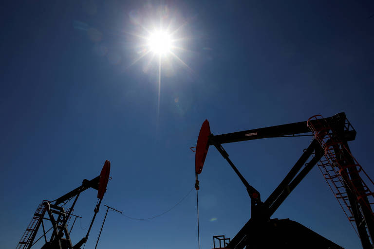 A imagem mostra silhuetas de plataformas de perfuração de petróleo contra um céu azul claro， com o sol brilhando intensamente no centro superior da imagem.
