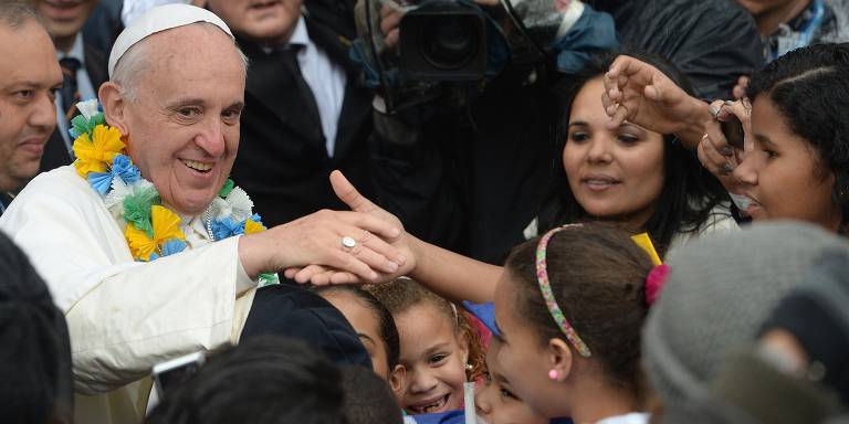 O papa Francisco está sorrindo e cumprimentando pessoas em uma multidão. Ele usa uma guirlanda de flores coloridas ao redor do pescoço e está estendendo a mão para cumprimentar uma criança. Ao seu redor, há várias pessoas, incluindo crianças e adultos, que estão tentando tocar ou cumprimentar o papa. O ambiente parece ser um evento ao ar livre, com algumas pessoas segurando câmeras.