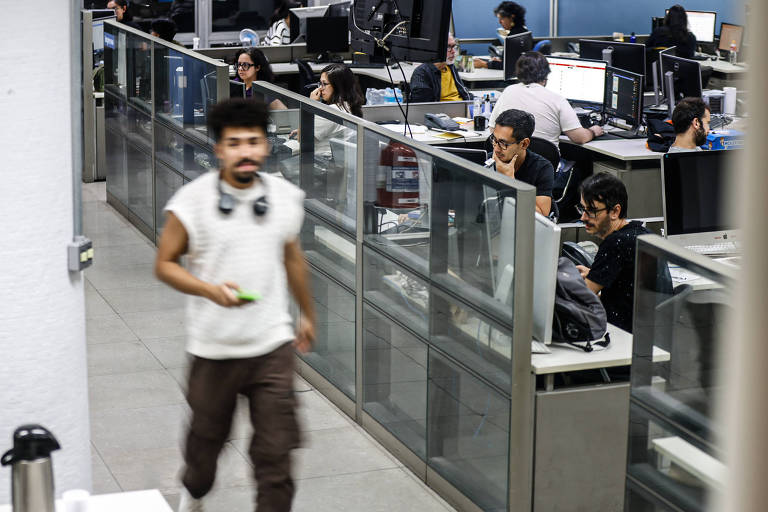 A imagem mostra um ambiente de escritório moderno， com várias mesas de trabalho. Algumas pessoas estão sentadas em frente a computadores， enquanto uma pessoa caminha em primeiro plano. As mesas são divididas por painéis de vidro. O ambiente é bem iluminado， com monitores e equipamentos de escritório visíveis.