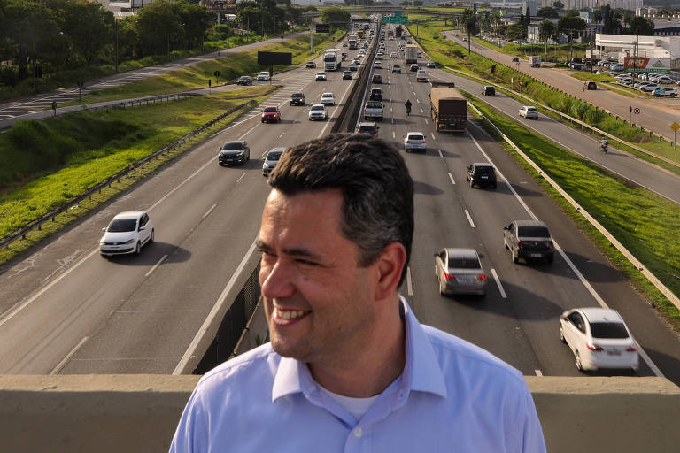 Um homem sorridente está posicionado em uma ponte， com uma rodovia movimentada ao fundo. A rodovia possui várias faixas de tráfego， com carros e caminhões se deslocando em direções opostas. O ambiente é urbano， com vegetação nas laterais e edifícios visíveis ao fundo.