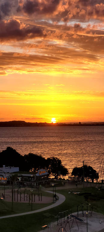 A imagem mostra um pôr do sol vibrante sobre um lago, com nuvens em tons de laranja e amarelo. No primeiro plano, há um parque com equipamentos de ginástica e um restaurante chamado 'DE RUA'. A luz do sol reflete na água, criando um efeito dourado. Ao fundo, é possível ver uma linha de árvores e edificações ao longo da costa.