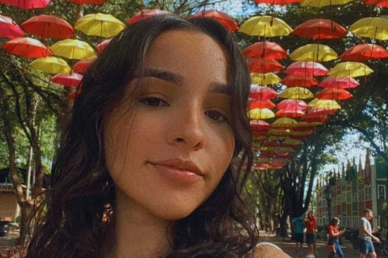 A imagem mostra uma jovem mulher tirando uma selfie em um ambiente ao ar livre， com guarda-chuvas vermelhos e amarelos pendurados acima dela. O fundo é composto por árvores e outras pessoas visíveis ao longe. A mulher tem cabelo ondulado e usa uma blusa branca.