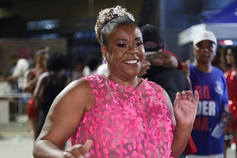 Uma mulher sorridente está dançando em um ambiente festivo. Ela usa um vestido rosa com detalhes em flores e uma saia volumosa. A mulher tem cabelo curto e usa uma tiara. Ao fundo, há outras pessoas e uma iluminação que sugere um evento noturno.