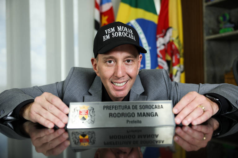 Um homem sorridente está sentado à mesa， usando um boné preto com a frase 039;VEM MORAR EM SOROCABA039;. Ele segura uma placa com o texto 039;PREFEITURA DE SOROCABA RODRIGO MANGA Prefeito039;. Ao fundo， há bandeiras e um ambiente de escritório.