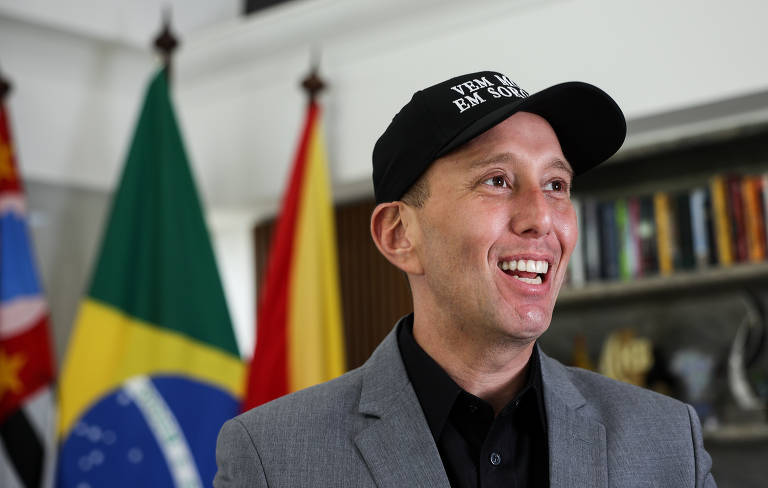 Um homem sorridente está usando um boné preto com a frase 'VEM MORAR EM SOROCABA'. Ele veste um paletó cinza e está em um ambiente interno, com bandeiras do Brasil, de São Paulo e de Sorocaba ao fundo. Há prateleiras com livros atrás dele.