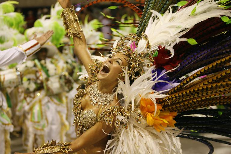 Uma dançarina de carnaval está em destaque, com um traje exuberante adornado com penas brancas, flores coloridas e muitos brilhos. Ela está com os braços levantados e a boca aberta, expressando alegria e entusiasmo. Ao fundo, outros dançarinos em trajes brancos e verdes estão visíveis, criando um ambiente festivo.