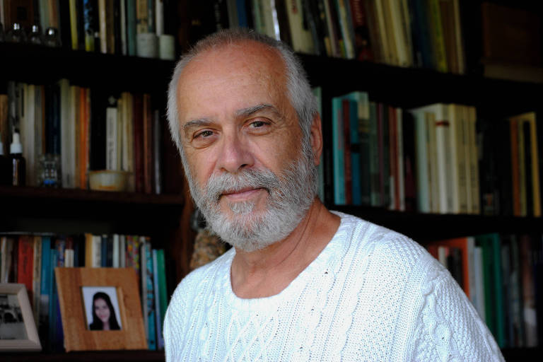 Um homem de cabelos grisalhos e barba， vestindo um suéter branco de tricô， está posando para a foto. Ele tem uma expressão serena e está olhando diretamente para a câmera. Ao fundo， há uma estante cheia de livros e uma pequena foto emoldurada de uma mulher.