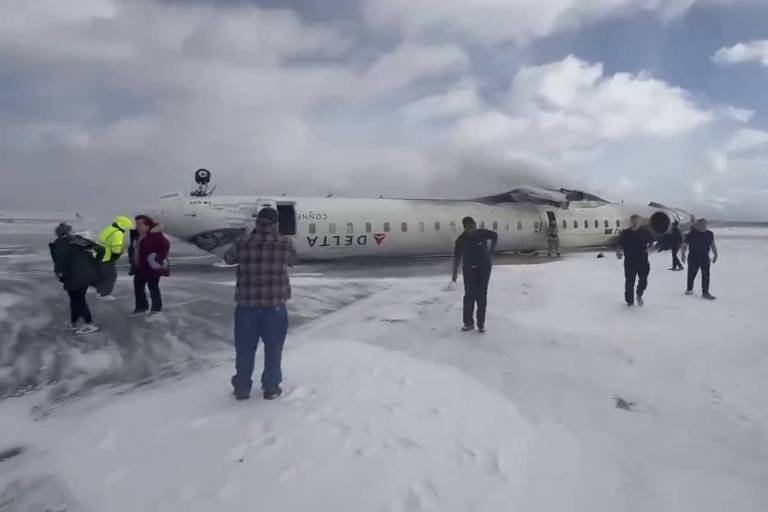 A imagem mostra um avião acidentado em uma pista coberta de neve. O avião， que parece ser um modelo de jato comercial， está parcialmente danificado e com fumaça saindo de uma de suas turbinas. Há várias pessoas ao redor do avião， algumas parecendo estar em estado de evacuação， enquanto outras estão se aproximando. Ao fundo， é possível ver um veículo de emergência.