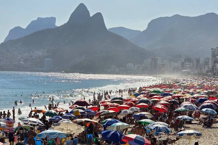 A imagem mostra uma praia lotada com muitas pessoas e guarda-sóis coloridos. Ao fundo， há montanhas com contornos distintos e uma cidade à direita. O céu está claro e o mar reflete a luz do sol.