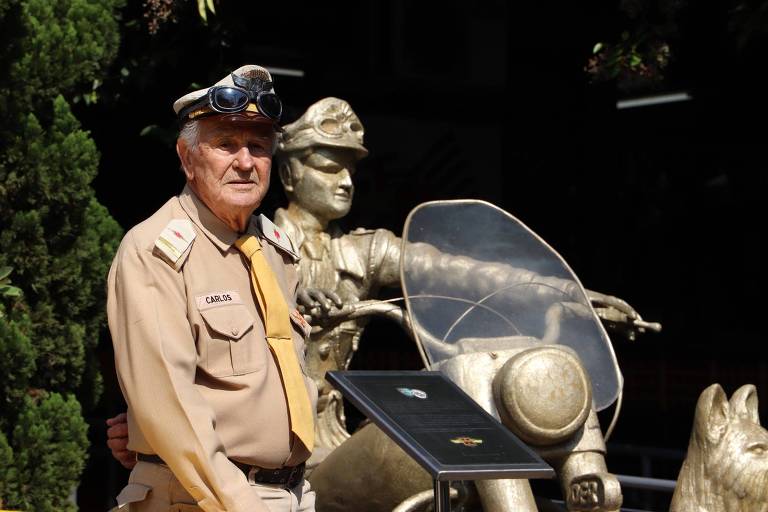 Um homem idoso， vestido com um uniforme de policial， posa ao lado de uma estátua de bronze de um motociclista. O homem usa um chapéu com óculos de proteção e está em frente a uma placa preta que parece conter informações. Ao fundo， há vegetação.