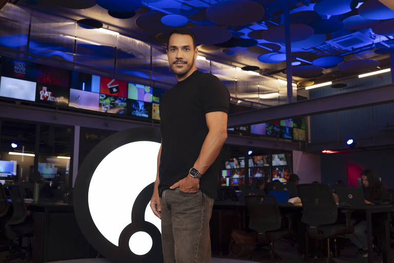 Um homem está posando em um estúdio de televisão. Ele usa uma camiseta preta e calças claras, e está de pé ao lado de um grande logotipo circular. Ao fundo, há várias telas exibindo conteúdo, e o ambiente é iluminado com luzes azuis e roxas.