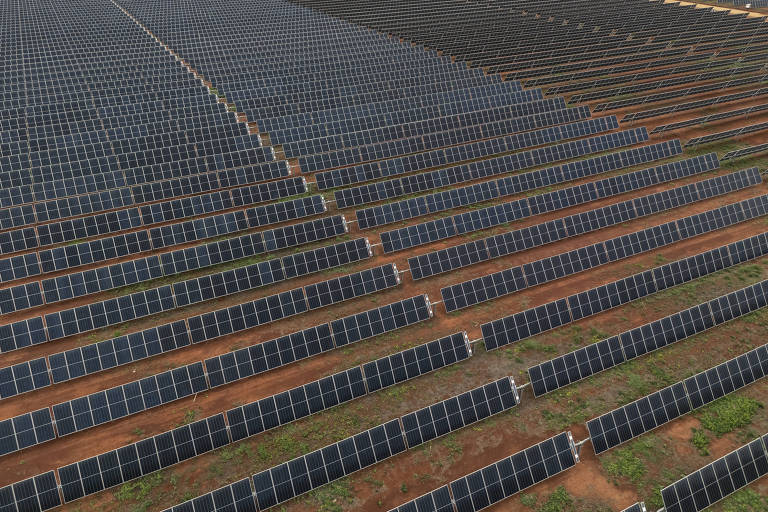 A imagem mostra um grande campo de painéis solares dispostos em linhas retas， cobrindo uma vasta área de terra. Os painéis são predominantemente pretos e estão alinhados de forma organizada， com espaços entre eles. O solo é visível entre as fileiras de painéis， apresentando uma coloração marrom e algumas áreas de vegetação.