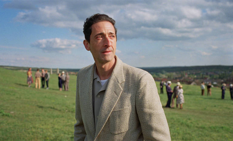 Um homem está em um campo verde, olhando para o horizonte. Ele usa um paletó claro e uma camiseta. Ao fundo, há várias pessoas em um grupo, algumas delas vestindo roupas coloridas. O céu está parcialmente nublado, com algumas nuvens visíveis.