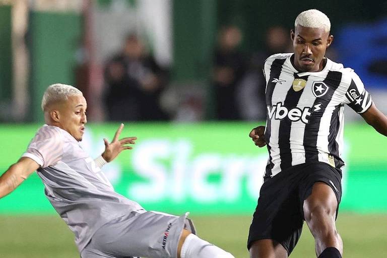 A imagem mostra dois jogadores de futebol em um momento de disputa. Um jogador está caindo no chão， enquanto o outro está em pé， com a bola próxima. O jogador à esquerda veste uma camisa clara e o jogador à direita usa uma camisa listrada em preto e branco. O fundo é desfocado， mas há uma iluminação que destaca os jogadores.