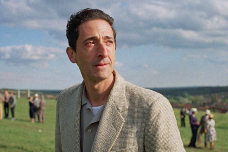 Um homem está em um campo verde， olhando para o horizonte. Ele usa um paletó claro e uma camiseta. Ao fundo， há várias pessoas em um grupo， algumas delas vestindo roupas coloridas. O céu está parcialmente nublado， com algumas nuvens visíveis.