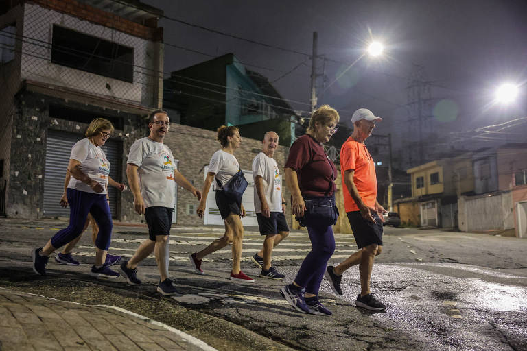 Um grupo de seis pessoas caminha em uma rua à noite. Elas estão vestindo camisetas brancas com um logo colorido e calças escuras. O ambiente é urbano, com casas e postes de luz ao fundo. O céu está escuro e há iluminação artificial nas ruas.