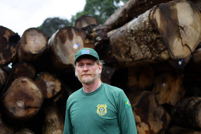 Um homem de cabelo comprido e barba， vestindo uma camiseta verde e um boné azul， está posicionado em frente a um grande empilhamento de toras de madeira. O fundo é composto por troncos de árvores empilhados， com algumas marcas visíveis na madeira. O ambiente parece ser uma área de floresta ou um local de armazenamento de madeira.