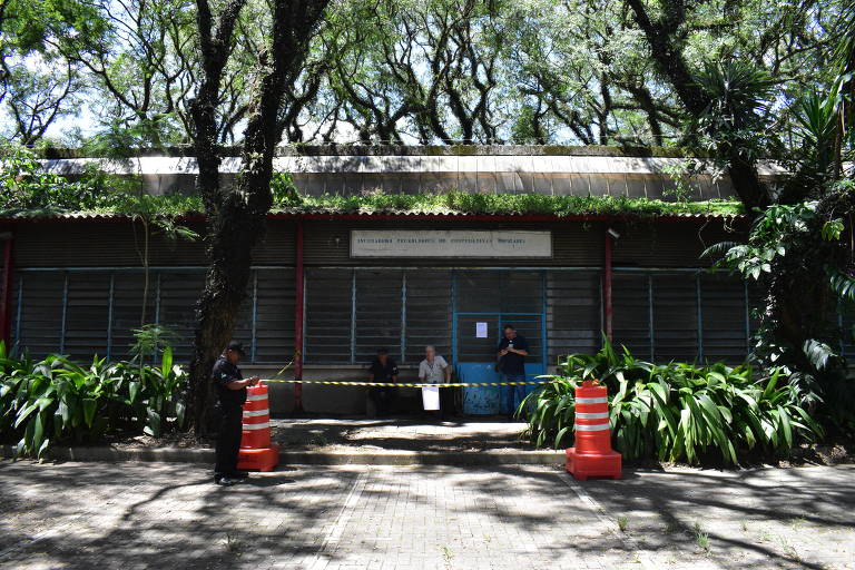 A imagem mostra a fachada de um edifício com janelas de metal fechadas e uma porta azul. Há pessoas sentadas em frente ao edifício， e a área está cercada por cones laranjas. O local é cercado por árvores e plantas verdes， criando um ambiente natural ao redor.