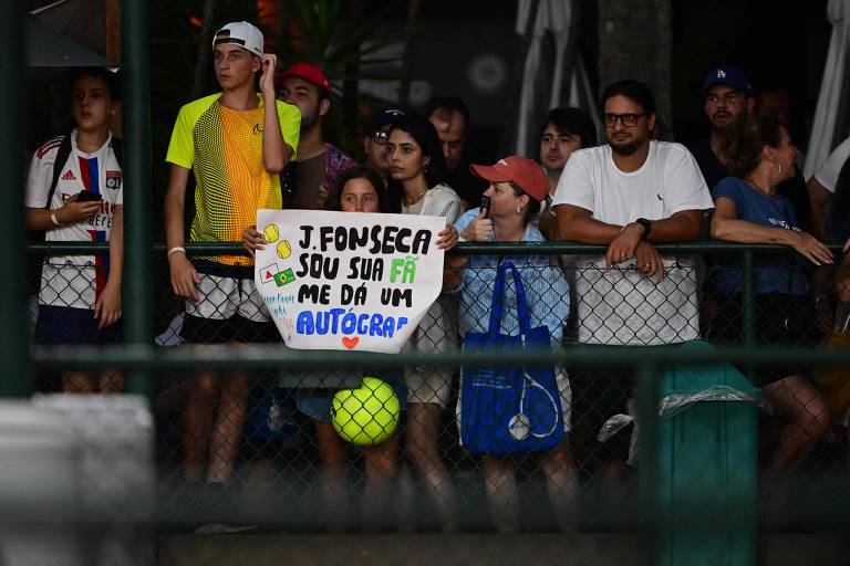 Um grupo de pessoas assistindo a um evento esportivo， com destaque para uma jovem segurando um cartaz que diz: 039;J. FONSECA SOU SUA FÃ ME DÁ UM AUTÓGRAFO039;. As pessoas estão em um ambiente ao ar livre， cercadas por uma grade， e algumas usam roupas esportivas.