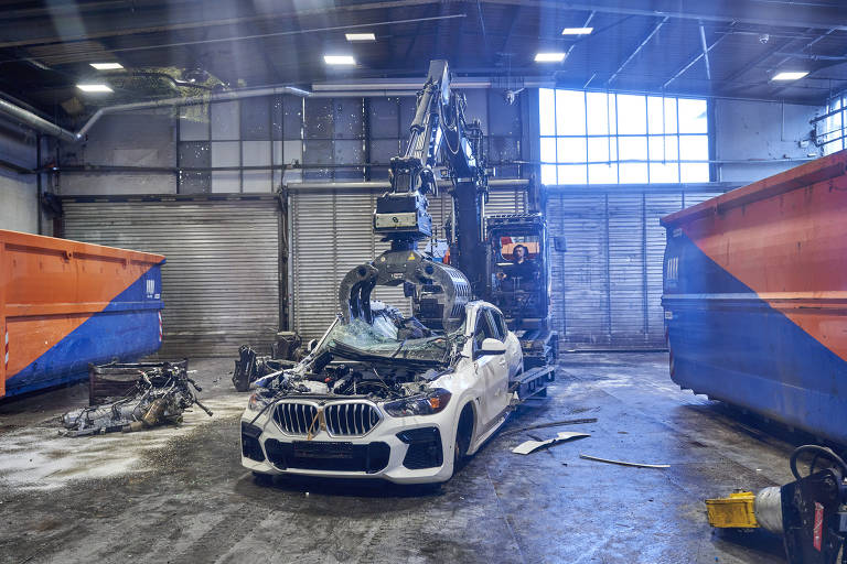 A imagem mostra uma oficina onde um carro BMW branco está sendo desmantelado. Um equipamento mecânico， semelhante a um braço robótico， está posicionado sobre o carro， aparentemente pronto para remover partes dele. Ao fundo， há janelas grandes que permitem a entrada de luz natural， e dois contêineres de lixo laranja e azul estão visíveis nas laterais. O chão parece sujo， com peças de carro espalhadas.