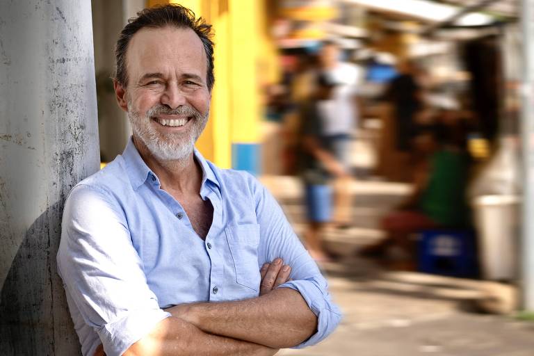 Um homem de meia-idade， com cabelo grisalho e barba， está sorrindo enquanto se apoia em um poste. Ele usa uma camisa de manga longa azul clara. Ao fundo， há um ambiente urbano desfocado， com pessoas e estruturas visíveis， sugerindo uma cena movimentada.