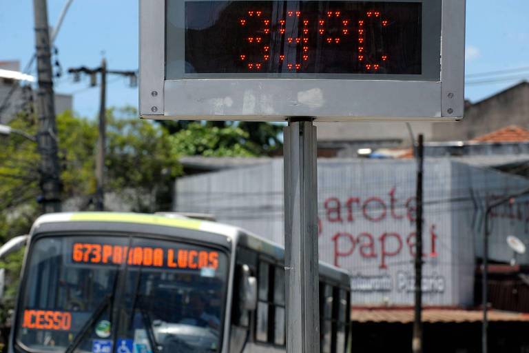 A imagem mostra um painel digital que indica a temperatura de 39°C. Em primeiro plano， há um ônibus com o número da linha 673， que vai para Pindamonhangaba. Ao fundo， é possível ver um comércio com a inscrição 039;Carota Papai039;.