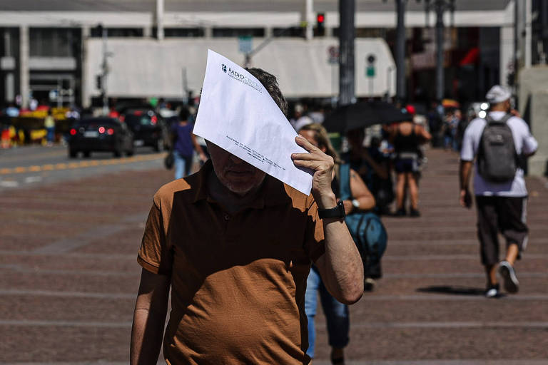 Um homem está caminhando em uma rua movimentada， segurando uma folha de papel sobre a cabeça， possivelmente para se proteger do sol. Ele usa uma camisa marrom e está em uma área com várias pessoas ao fundo， algumas com guarda-sóis. Há carros passando na rua ao fundo.