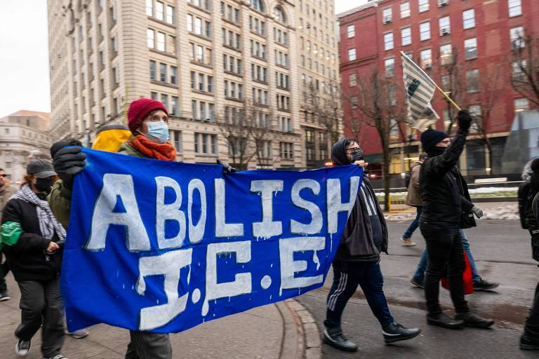 Um grupo de manifestantes caminha em uma rua， segurando um grande banner azul que diz 039;acabe com a ICE039;， a sigla para a agência de imigração americana， em letras brancas. Algumas pessoas usam máscaras e estão vestidas de forma variada， enquanto outras seguram bandeiras. O cenário urbano inclui prédios altos ao fundo e árvores sem folhas.