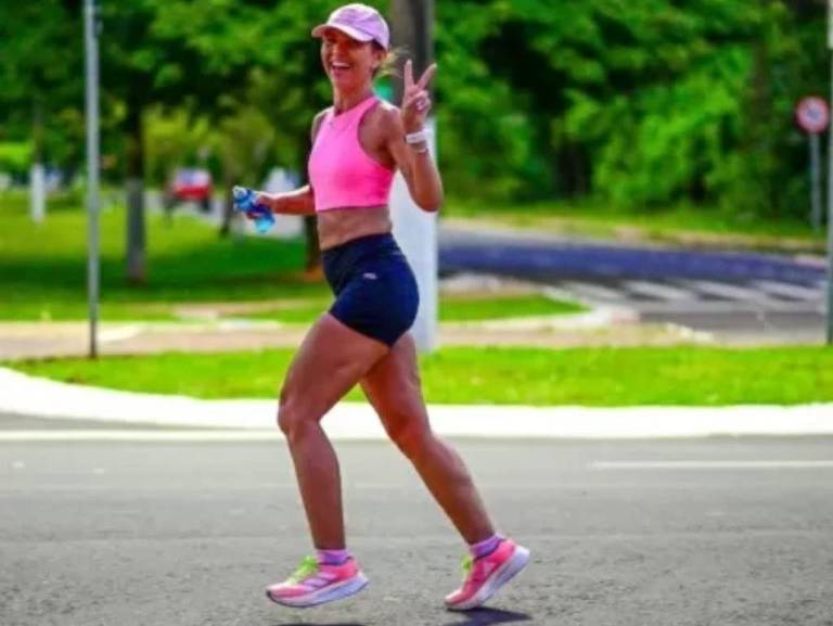 Uma mulher está correndo em uma estrada， vestindo um top rosa e shorts escuros. Ela sorri e faz um sinal de paz com a mão. Ao fundo， há uma área verde e uma estrada pavimentada.