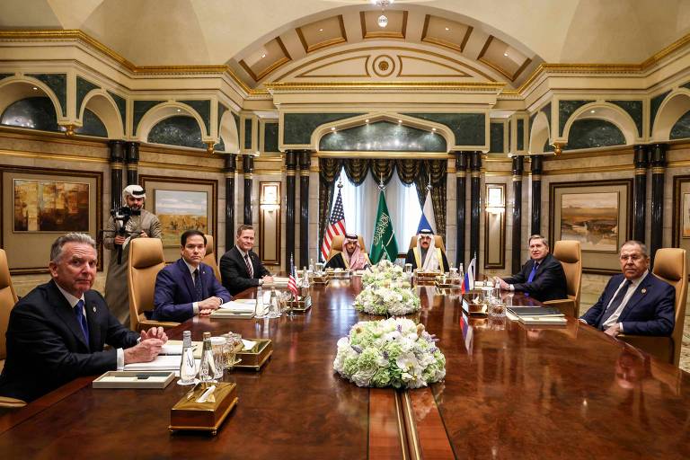 A imagem mostra uma reunião em uma sala de conferências luxuosa. Há uma mesa grande em formato retangular com várias pessoas sentadas ao redor. No centro da mesa， há um arranjo de flores. Ao fundo， há cortinas brancas e uma bandeira dos Estados Unidos， uma bandeira da Arábia Saudita e outras bandeiras visíveis. As pessoas estão vestidas formalmente， e um homem em pé ao fundo parece ser um segurança.