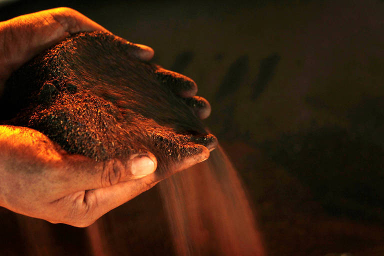 Uma mão segurando uma quantidade de areia ou terra， com grãos caindo entre os dedos. A iluminação é suave， destacando a textura da areia e a cor avermelhada do material. O fundo é escuro， o que faz com que a mão e a areia se destaquem.