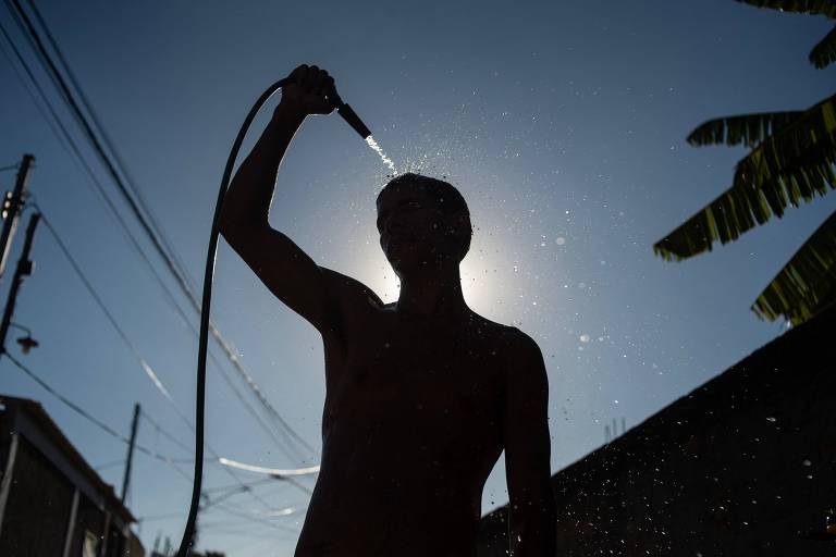 A imagem mostra a silhueta de um jovem sem camisa segurando uma mangueira de água， enquanto a água é jorrada sobre sua cabeça. O fundo é iluminado pelo sol， criando um efeito de contraluz. Há fios elétricos visíveis e uma planta ao lado， sugerindo um ambiente externo.