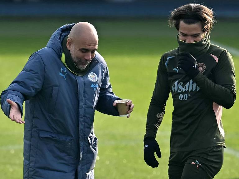 A imagem mostra um treinador， que é Pep Guardiola， e um jogador de futebol， que é Jack Grealish， durante um treino do Manchester City. O treinador， vestido com um casaco azul e segurando um copo， parece estar conversando com o jogador， que está usando um agasalho verde escuro e cobrindo parte do rosto com um cachecol. O cenário é um campo de futebol com grama verde ao fundo.