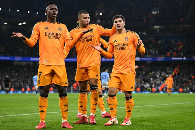 Em partida contra o Manchester City， três jogadores do Real Madrid estão em campo， vestindo uniformes da cor laranja com o logo da equipe e a inscrição 039;Emirates Fly Better039;. Eles são Vinicius Junior， Mbappé e Brahim Díaz e estão em uma pose descontraída， com expressões de confusão ou indagação. O fundo mostra um estádio de futebol com torcedores.