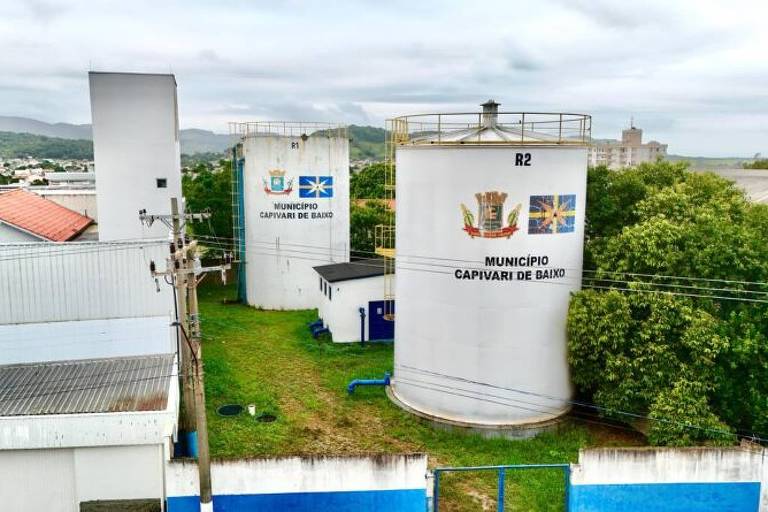 A imagem mostra tanques de armazenamento em um ambiente externo. Os tanques são de formato cilíndrico e estão localizados em uma área verde， com algumas árvores ao redor. Ao fundo， há uma construção de telhado vermelho e uma paisagem urbana visível. O céu está nublado.