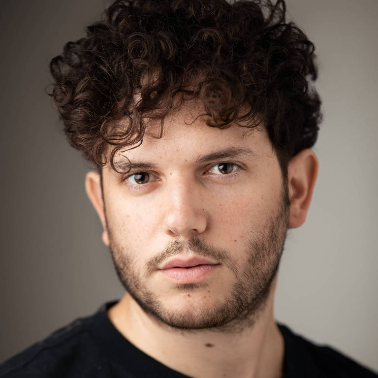 Um homem com cabelo cacheado e barba， olhando diretamente para a câmera. Ele está vestindo uma camiseta preta e o fundo é neutro， em tons claros.