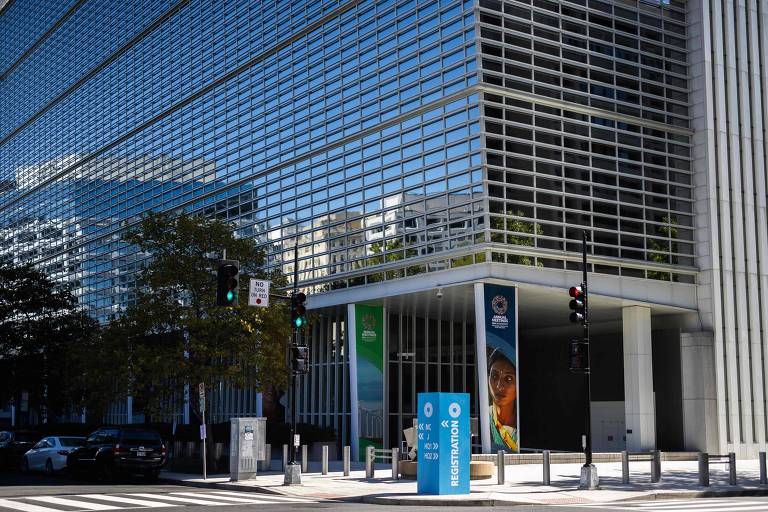 A imagem mostra a fachada do Banco Mundial， um edifício moderno com uma grande quantidade de janelas de vidro. À esquerda， há árvores e um sinal de trânsito. Na entrada do edifício， há um painel com a imagem de uma mulher e um letreiro azul com informações. O dia parece ensolarado.