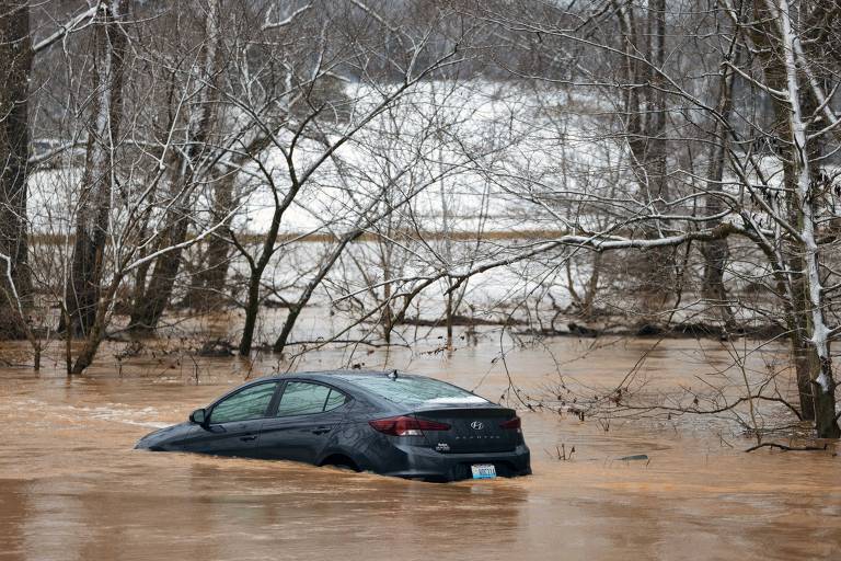 A imagem mostra um carro preto parcialmente submerso em água de enchente， cercado por árvores sem folhas. O cenário é de um ambiente rural， com um fundo de neve visível no solo. A água é de cor marrom， indicando a presença de lama e detritos.