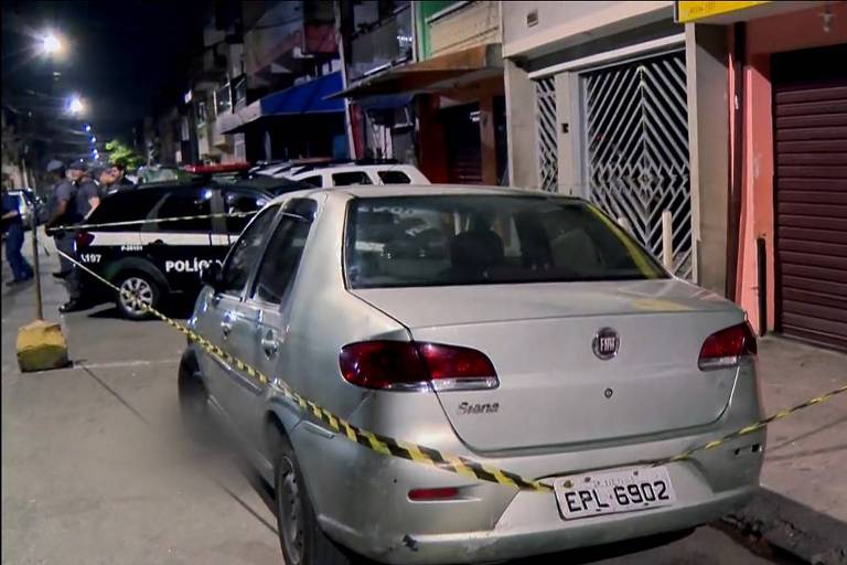 A imagem mostra uma rua à noite com um carro prata estacionado em primeiro plano. O veículo está cercado por fitas de isolamento amarelas e pretas. Ao fundo， há uma viatura da polícia e algumas lojas com fachadas coloridas. Uma das lojas tem uma porta de metal fechada e há cartazes colados na parede ao lado. A iluminação é fraca， típica de uma cena noturna.