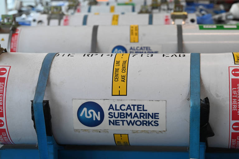 A imagem mostra vários cilindros brancos dispostos em uma linha， com etiquetas coloridas e faixas de segurança. Um dos cilindros possui o logotipo da Alcatel Submarine Networks e a inscrição 039;CENTRE USE AXE CENTRAL039;.