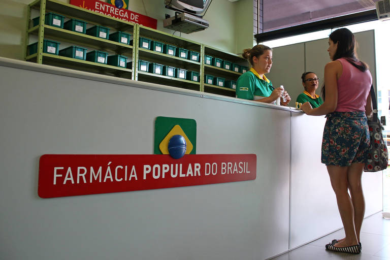 A imagem mostra o interior de uma farmácia popular no Brasil. No balcão， uma mulher está conversando com duas funcionárias que usam uniformes verdes. Ao fundo， há prateleiras com caixas organizadas. Na parede， há um letreiro vermelho com o texto 039;FARMÁCIA POPULAR DO BRASIL039; e um símbolo da bandeira do Brasil.