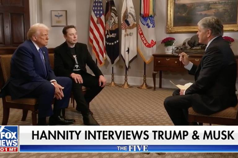 A imagem mostra uma entrevista realizada por Sean Hannity， onde ele está sentado em uma cadeira， segurando um bloco de notas. À sua esquerda， está Donald Trump， vestido com um terno azul e uma gravata vermelha. À direita， Elon Musk， vestido com uma camiseta preta e calças escuras. Ao fundo， há bandeiras e um quadro na parede， indicando um ambiente formal.