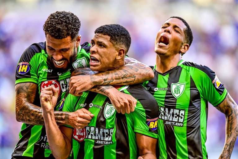 Três jogadores de futebol estão se abraçando e comemorando em um campo. Eles vestem uniformes verdes e pretos， com detalhes em branco. O jogador do meio parece estar gritando de alegria， enquanto os outros dois estão sorrindo e demonstrando entusiasmo. O fundo é desfocado， sugerindo um estádio cheio.