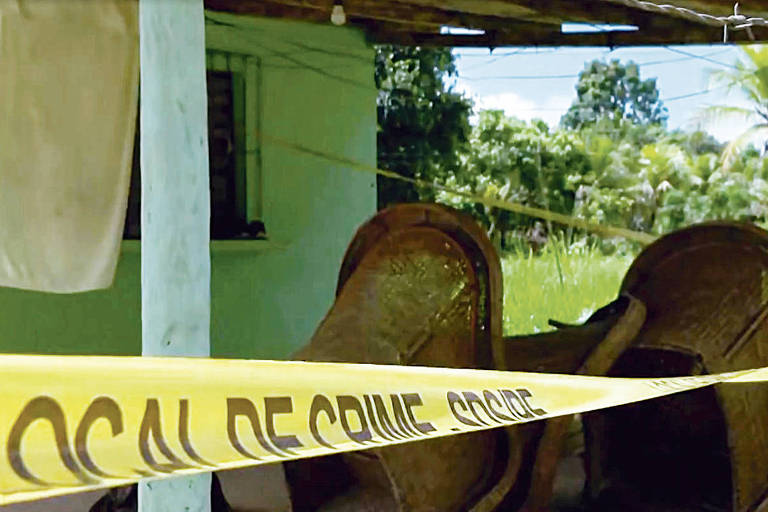 A imagem mostra uma área externa de uma casa com uma fita amarela de 039;LOCAL DE CRIME039; estendida. Ao fundo， há uma parede verde e janelas com grades. Dois sofás estão virados de lado， e há um pano pendurado em um varal. O ambiente é cercado por vegetação.