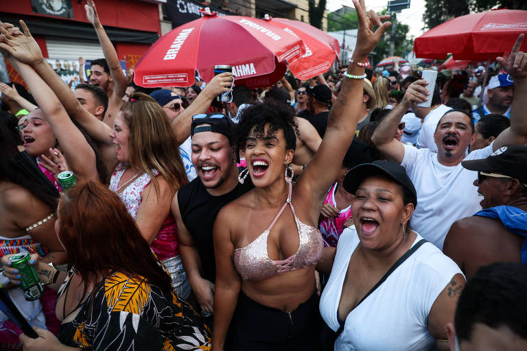 Uma multidão animada participa de uma festa de rua， com pessoas sorrindo e levantando as mãos. Algumas pessoas seguram garrafas de bebida e há guarda-sóis vermelhos ao fundo. A atmosfera é de celebração e alegria.